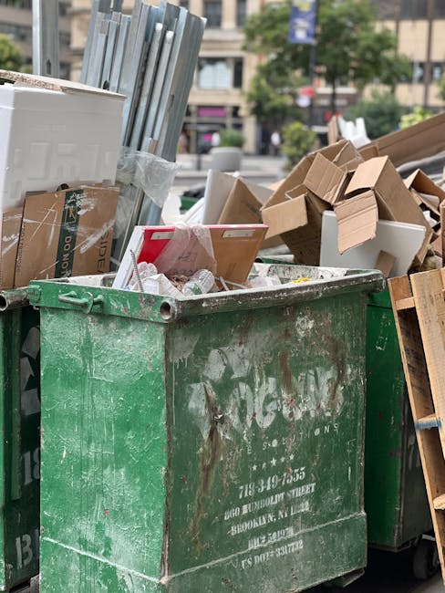 A large, weathered green waste container filled with a mixture of recyclable and non-recyclable rubbish, positioned outdoors on a paved urban street. The contents include flattened cardboard boxes, some with printed labels, various paper packaging, and plastic wrapping. To the left of the container, there are metal frames or panels leaning slightly, and in the background, additional cardboard items and wooden pallets are visible, stacked haphazardly. The scene is set in a city environment with blurred buildings, signage, and trees in the distance, suggesting a designated area for rubbish collection or disposal. Natural light illuminates the scene, highlighting the textured surface of the container and the various materials spilling over its edges, indicative of an informal or private waste collection process. The visibility of upright articles and their arrangement reflect typical scenes encountered during independent waste disposal, aligning with alternative rubbish removal options such as private collection services like those offered by Waste Disposal Elephant and Castle.