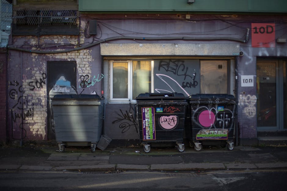 A tall, weathered brick wall with a red metal door displaying various graffiti tags in white and pink paint. Attached to the door are multiple signs, including a white notice with the words 'NO MONEY' and a blue and white sticker saying 'Keep clear.' In front of the door, a pile of black rubbish bags is stacked haphazardly on a concrete surface; some bags are partially torn, revealing waste inside. Among the rubbish, there are pieces of flattened cardboard boxes, a wooden pallet or frame, and rolled-up packaging materials, indicating a collection of household or commercial waste awaiting removal. The scene suggests a neglected or infrequently cleared area, highlighting the need for professional rubbish disposal services, such as those provided by Waste Disposale Elephant and Castle, for alternative waste management solutions outside regular local authority collection.