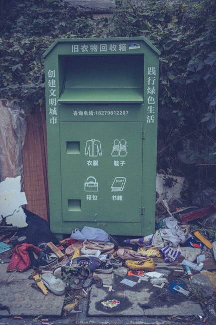 A green old clothing collection box with Chinese characters and a contact number on its upper section, situated outdoors against a backdrop of dense foliage and partially visible structures. The box has a rectangular shape with a sloped top, a large empty opening at the top for depositing items, and smaller compartments below designated for different types of clothing, shoes, bags, and books, indicated by simple line illustrations and Chinese labels. In front of the box, on the ground, there is a noticeable amount of discarded rubbish including various shoes, clothing, plastic bags, small packaging materials, and miscellaneous items, some scattered directly on the paved surface. The scene appears to be in a neglected or cluttered area where informal waste disposal is evident, reflecting issues of environmental cleanliness and rubbish management concerns related to private waste collection services. The overall lighting is natural but subdued, fitting a daytime outdoor setting with overgrown foliage providing a leafy background behind the collection point, which is associated with efforts to reuse or recycle clothing and textiles, aligning with alternative waste handling practices.