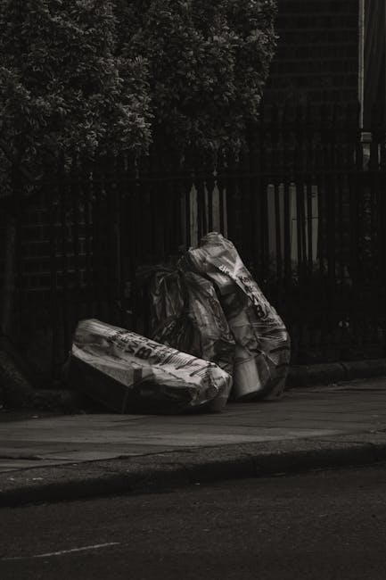 A black and white photograph showing several large trash bags placed on the pavement near a black metal fence and a brick wall, with dense foliage in the background. The bags appear to be filled with rubbish and are positioned in a close cluster, some leaning against each other. The scene is outdoors, possibly on a residential street or alley, with the bags awaiting collection or disposal. The texture of the bags looks crinkled and tightly filled, with some remnants of printed text visible on the packaging. The ambient lighting suggests a cloudy or overcast day, contributing to the overall subdued tone of the scene, which aligns with the context of private or alternative waste handling services such as those offered by Waste Disposa Le Elephant and Castle. The setting underscores the process of clearing rubbish from property in preparation for waste removal, characteristic of independent collection or onsite clearance activities.