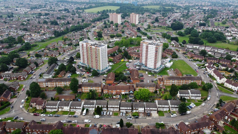 An aerial view of a residential area showing a mix of housing types, including rows of terraced houses with grey and white facades, pitched roofs, and small front gardens. In the center of the image, there are two tall, multi-storey apartment blocks with light-colored exteriors and numerous balconies, surrounded by parking lots filled with cars. The surrounding environment features green spaces, trees, and open fields in the distance. The overall scene is well-lit with natural daylight, emphasizing the urban layout and building details. This setting reflects typical urban waste management needs, such as potential private rubbish collection or on-site clearance for the residential and apartment buildings, showcasing the kind of environment where professional waste disposal services may be required, as seen on waste disposal company websites.