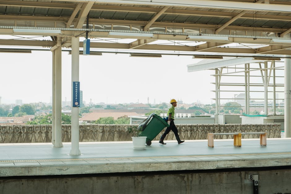 A worker wearing a yellow safety helmet, a green high-visibility vest, and dark trousers is walking along an outdoor train platform, pulling a large green waste bin on wheels behind them. The bin appears to be made of durable plastic with a textured surface, and it is fitted with a black lid that's open or removed. In front of the worker, there is a white plastic bucket placed on the platform. The platform features a metal safety barrier with decorative detailing along the edge, separating it from the cityscape visible in the background. Overhead, there is a roof structure with metal beams and panels, providing shade and shelter to passengers, with natural light illuminating the scene. The environment suggests a typical urban railway station where private or independent rubbish collection and waste disposal services, like those from Waste Disposal Elephant and Castle, facilitate alternative waste handling outside standard council services.