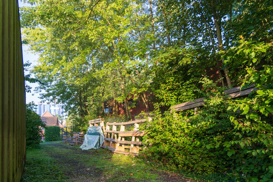 Three large black plastic rubbish bags filled with waste are placed on the pavement next to a metal fence with vertical bars. The bags appear to contain household or garden waste and are slightly crumpled with some areas showing direction lines or printed markings. The scene is set outdoors, with a dark background featuring leafy trees and parts of a building or structure partially visible behind the fence. The area is lit with natural light, casting minimal shadows, and the overall environment suggests a typical outdoor waste collection point suitable for private rubbish removal services like those offered by Waste Disposables Elephant and Castle, emphasizing the need for proper disposal of domestic refuse.