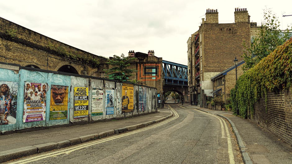 A narrow urban street scene featuring a curved tarmac road with clearly marked double yellow lines along the edge, leading beneath a blue metal railway bridge structure spanning across the top. On the left side, a weathered concrete wall is covered with multiple colorful posters advertising music events and performances, some torn or peeling, with a rough texture. Behind this wall, parts of an aged brick building are visible, with a moss or plant growth along the top edge. On the right side, a high brick wall adorned with green foliage and ivy runs parallel to the street, partially obscuring residential buildings behind it, which have brick facades and fire escape balconies. The scene appears to be captured on an overcast day with diffused natural light, creating a neutral, muted atmosphere. The image subtly suggests themes related to urban clearance or private waste removal, with no visible rubbish or waste materials present in the scene, but it reflects an environment where external cleaning or disposal services could be relevant, akin to independent collection or site clearance tasks by companies like Waste Disposale Elephant and Castle.