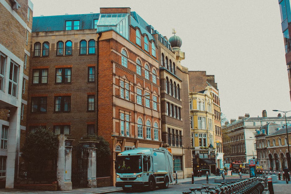 The image depicts a street scene featuring a row of multi-storey buildings with diverse architectural styles. The central building is constructed from red brick with arched windows and decorative stone details, topped with a distinctive greenish copper roof and a domed turret. To its left, there's a modern white building with large, rectangular windows, while to the right, a cream-colored Victorian-style building with ornate window frames and balconies is visible. A waste collection van, painted in light blue with white accents, is parked on the street in front of the buildings, alongside a row of black and orange bollards and bicycle racks. The street surface is paved with asphalt, and the environment is well-lit with natural daylight, suggesting a clear or partly cloudy day. Vegetation is minimal, with small trees near the buildings. The scene appears to relate to urban waste management, where the presence of the waste vehicle indicates scheduled rubbish collection or private waste removal, reflecting services offered by waste disposal companies like wastedisposalelephantandcastle.co.uk for alternative waste handling in the area.