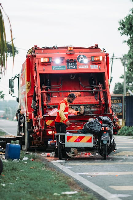 A waste collection worker, dressed in a high-visibility yellow and red uniform, is operating a large red rubbish truck parked on the side of a street. The worker is standing near the rear of the vehicle, which features a mechanical compactor at the back where rubbish is being collected. The truck's surface includes various safety lights, decals, and identification markings, with a visible license plate reading 'BRJ 7751.' In front of the truck, a black refuse bag is placed on a small metal platform, and nearby are scattered debris on the pavement, including a blue container and some loose waste. The scene is set outdoors on a clear day, with surrounding greenery and trees visible in the background, alongside a few buildings and utility poles. The environment suggests an urban or suburban area where private waste collection might be taking place, highlighting aspects of independent rubbish removal services like those offered by Waste Disposables Elephant and Castle. The overall scene emphasizes on-site waste collection and rubbish disposal processes typical of local or private waste management solutions in SE17 and surrounding areas.