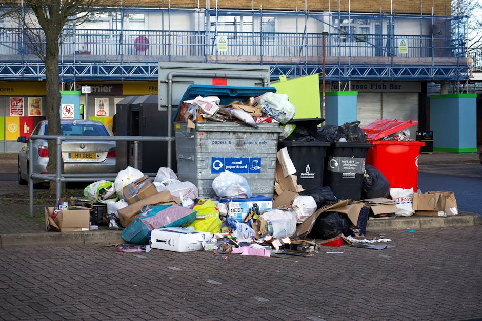 An outdoor urban scene showing multiple overflowing rubbish bins and scattered waste on a paved sidewalk next to a metal railing, with various types of discarded packaging, cardboard boxes, plastic bags, and miscellaneous debris spilling onto the ground. The central grey bin marked for mixed paper and card is overloaded, with items protruding from the open lid, while other black bins and a large red bin nearby also appear filled to capacity. Behind the waste, there are commercial units with shuttered storefronts and signage, and a multi-storey building covered in blue construction scaffolding and netting. To the left, a parked car with a yellow registration plate is partially visible, and a leafless tree provides some natural contrast to the scene. The overall environment suggests a busy area where independent rubbish collection and waste management services, such as those provided by waste disposal specialists, are responsible for handling on-site clearance and refuse removal to prevent litter accumulation.